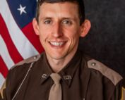 Portrait of a male sheriff in a brown uniform with a badge and name J. Samuelson, posed in front of the American flag.